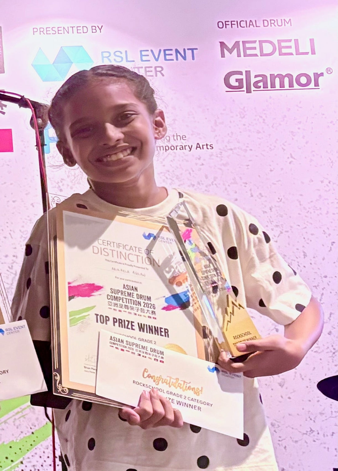 Mikaela Agilan holding Certificate of Distinction and Top Prize Gold Trophy for Rockschool Grade 2 at ASDC 2026 Singapore - daughter of Joshua Agilan Drumstick Studios Founder
